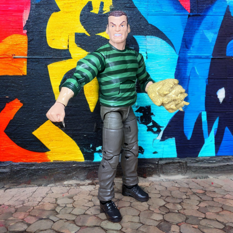 Les bras et les jambes sablonneux détaillés de la figurine Sandman 2025 avec des vêtements à rayures vertes, posés avec un accessoire en forme de poing sablonneux dans un présentoir de collection.