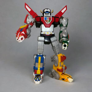 プレイメイトの5頭のライオンを合体させた16インチのボルトロンフィギュアは、その象徴的なフォルムを強調し、プレーンなグレーの背景に堂々と立っている。.