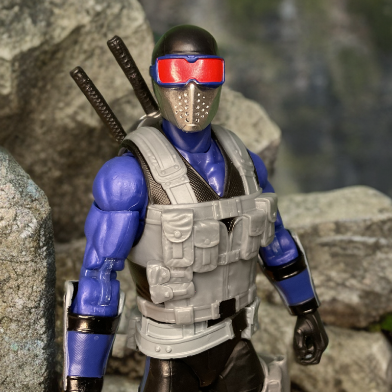 G.I. Joe Classified #165 Snake Eyes ist eine aufrecht stehende Figur mit einer Maschinenpistole in der Hand, einem blau-grauen Anzug, einer roten Schutzbrille und detaillierten Zubehörtaschen auf weißem Hintergrund.