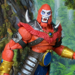 Uma visão detalhada da figura de ação New Eternia Beast Man do Masters of the Universe Masterverse, mostrando sua escultura de pelo laranja de 7 polegadas com pintura de guerra azul, mandíbula articulada e armadura removível com joias. A figura está em um cenário de selva rochosa, segurando um chicote azul metálico e uma maça com espinhos, com acessórios adicionais como um cocar com chifres e mãos extras. O design lembra o MOTU Origins Lords of Power Beast Man, combinando a estética de protótipo vintage com recursos modernos de colecionador.