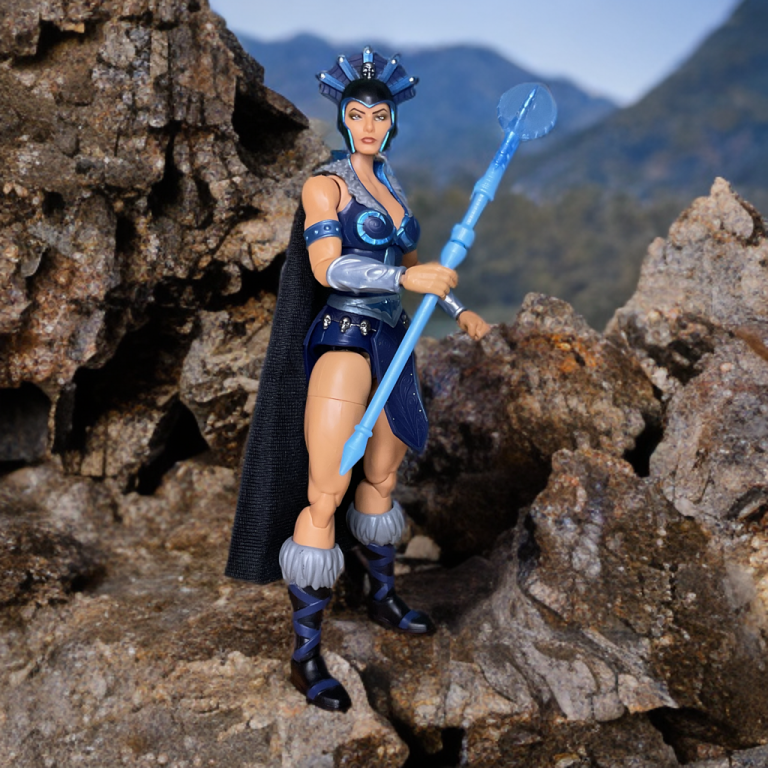 Die 7-Zoll New Eternia Evil-Lyn Actionfigur steht in einer dynamischen Pose da und präsentiert ihr blau-lila Gothic-Outfit, den Totenkopf-Helm und den im Dunkeln leuchtenden Stab vor einer Bergkulisse.