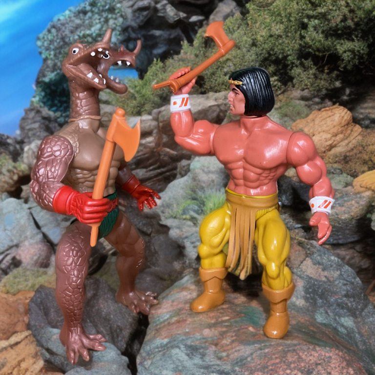Arak, con el hacha en la mano, se enfrenta a la figura bicéfala de Hydraz en un afloramiento rocoso bajo un cielo azul, parte de una sesión fotográfica que celebra la línea de juguetes Warlord de Remco de los años 80.