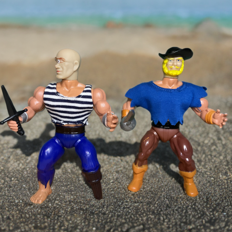 On a pebbled beach with a calm aqua horizon, Pegleg, a bald pirate with a glowing third eye on his forehead, stands in a striped black-and-white shirt, blue pants, and a peg leg, holding a dagger. Beside him, Cutlass, in a black hat and tattered blue tunic, grips a silver cutlass, his yellow beard flowing, with brown pants and orange boots.