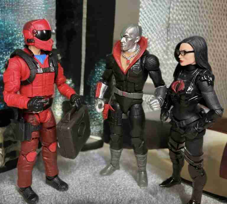 In a dimly lit industrial setting, Wild Weasel, in a red flight suit, hands a black briefcase to Destro, a metallic-skinned figure in a black uniform with a red collar. The Baroness, in a glossy black tactical outfit, observes intently, with metallic walls reflecting a faint blue glow.