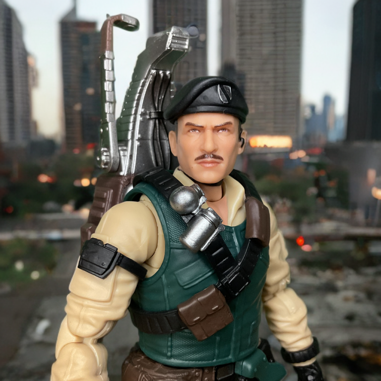 Dial-Tone’s sculpted face with mustache and shades, paired with his olive drab fatigues and detailed headset with a city in the background.