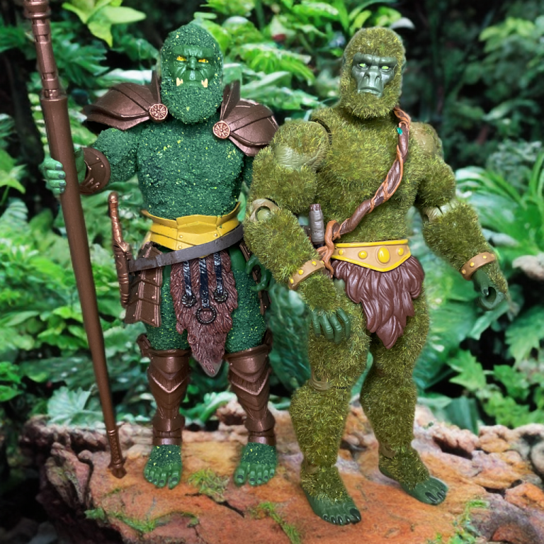 Bryophytus y el Hombre de Musgo están uno junto al otro sobre un tronco musgoso en un frondoso bosque. Bryophytus, a la izquierda, sostiene un largo bastón y lleva una detallada armadura de bronce con una característica textura musgosa que cubre su cuerpo. El Hombre de Musgo, a la derecha, está cubierto de un "musgo" verde suave y velloso y tiene un aspecto más simiesco, sosteniendo una rama frondosa sobre el hombro.
