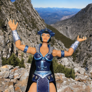 Neue Eternia Evil-Lyn-Actionfigur in einer zaubernden Pose, mit austauschbaren Händen, einem leuchtenden Kugelstab und einem wallenden Umhang vor einer Bergkulisse.