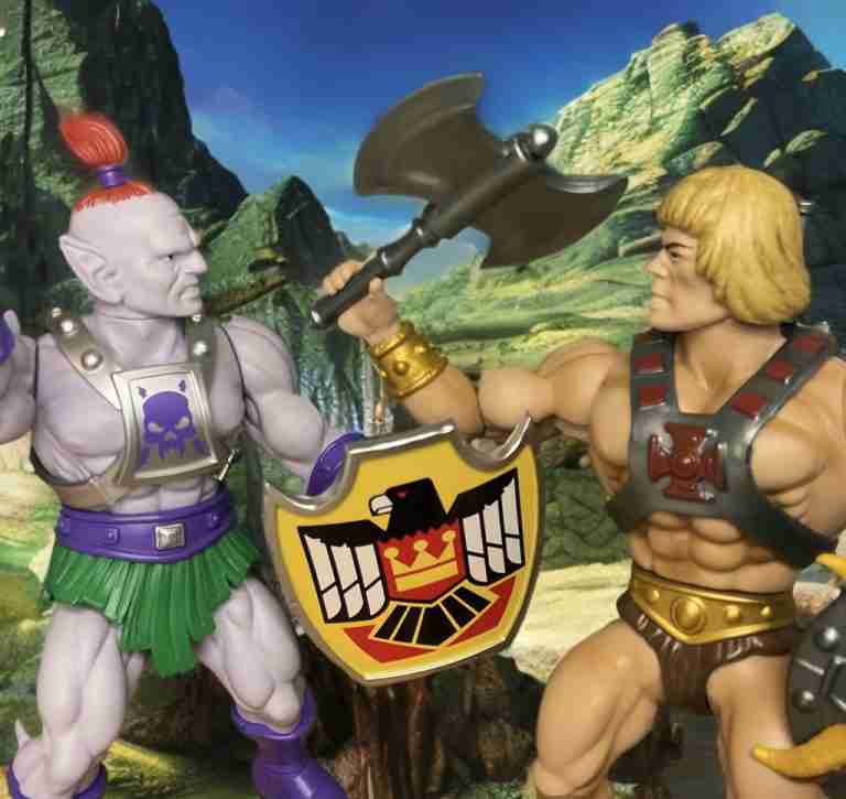 Heroic He-Man in battle harness facing off against Baltard in a verdant Eternian field, axe raised and shield blocking, with majestic mountains in the background.