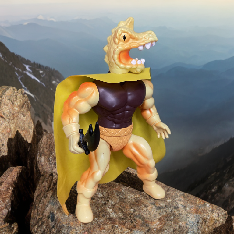 Ramyr, a figura amarela com cabeça de crocodilo, foice e capa amarela, posou em uma rocha com vista para montanhas enevoadas.