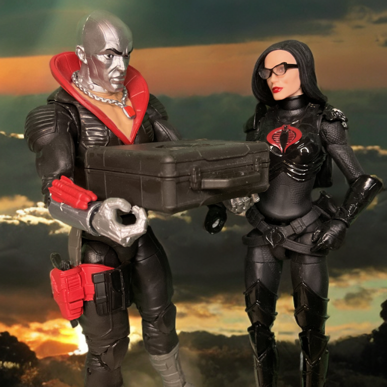 Destro, a metallic-skinned figure in a black uniform with a red collar, and the Baroness, in a glossy black tactical suit, stand on rugged terrain at sunset. Destro holds a black briefcase, both gazing at it under a vibrant orange and purple sky casting long shadows.