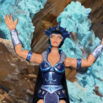 The New Eternia Evil-Lyn figure, with a sleek sculpt and metallic blue helmet, holds a glowing orb staff, displayed in collector-friendly packaging with vibrant Snake Mountain artwork.