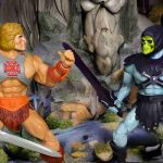 Un primer plano de la musculosa figura de He-Man, ataviado con un arnés y un taparrabos, empuñando su hacha, de pie sobre un saliente rocoso se defiende de la siniestra presencia de Skeletor.
