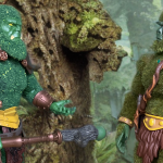 Against a backdrop of jagged, rocky cliffs and distant mountains, Bryophytus and Moss Man face each other. Bryophytus gestures with an open hand as if in conversation, while Moss Man listens intently, creating a sense of dialogue and shared purpose between the two figures.