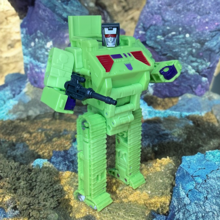 Bonecrusher bulldozer pushing forward with raised blade, grey-green camo paint gleaming in Hasbro’s Deluxe-class ’86 tribute.
