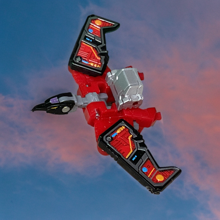 Red and black Laserbeak toy in bird mode, flying with wings outstretched, against a gradient blue-to-pink sky background.