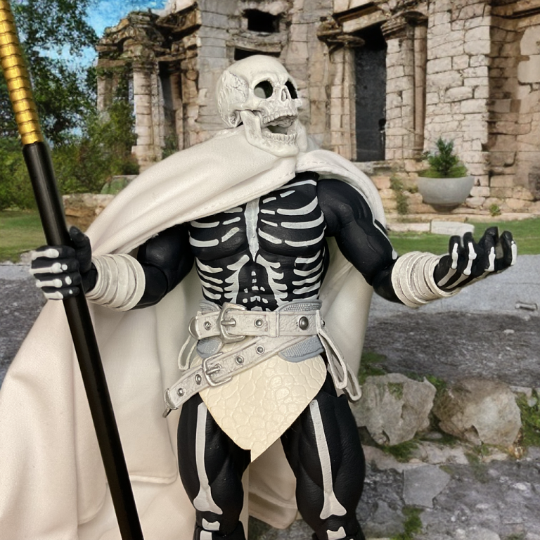 The Head Hunter figure in ancient ruins, wearing a white cape and holding a gold-tipped staff, its black body with white bones, white boots, and a silver belt, with stone columns and greenery in the background.