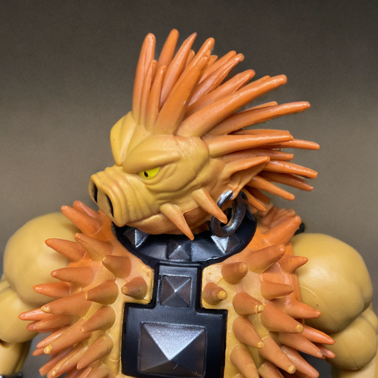 A close up of 5.5-inch Spikor figure with brown spikes and earrings. The packaging displays a black-and-white sketch of Spikor lunging, with orange accents and lore text.