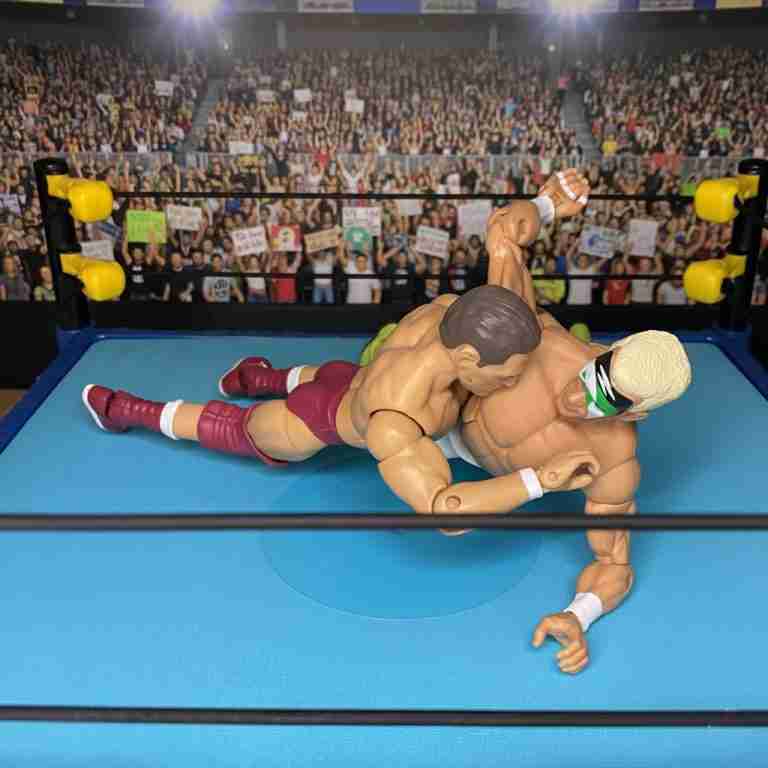 Nahaufnahme im Ring von Regal, der den Regal Stretch gegen einen ungenannten Gegner einsetzt, dessen Muskeln im Scheinwerferlicht der Arena angespannt sind.