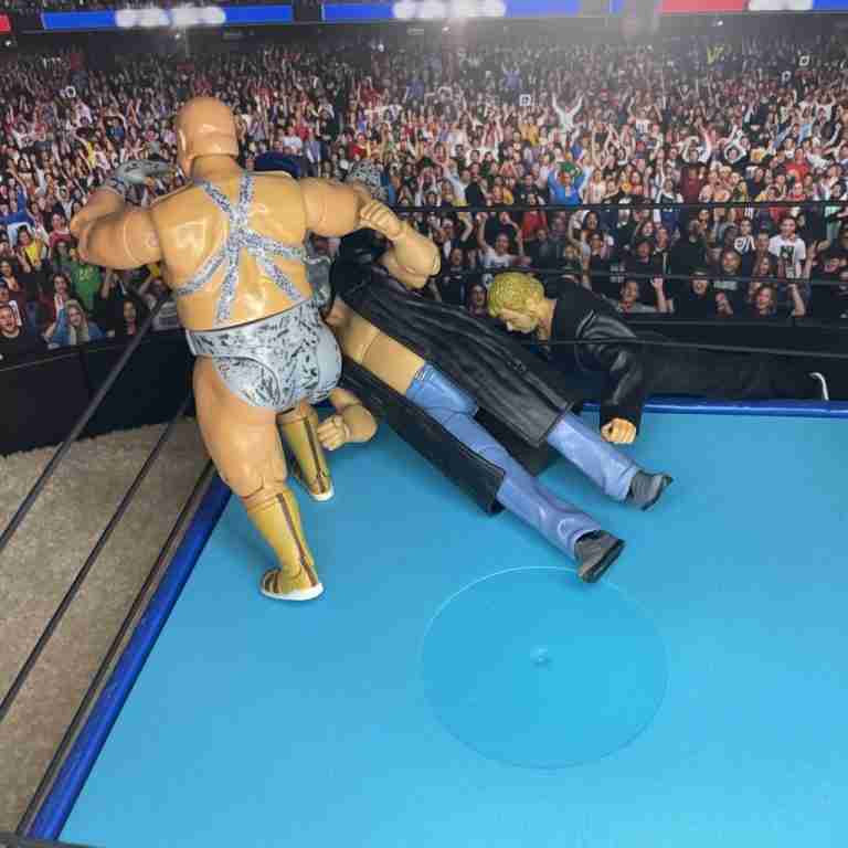 Die Actionfigur Bastion Booger stampft auf Shockmasters Kopf, während er auf dem obersten Seil steht und Race vom Ring aus anfeuert.