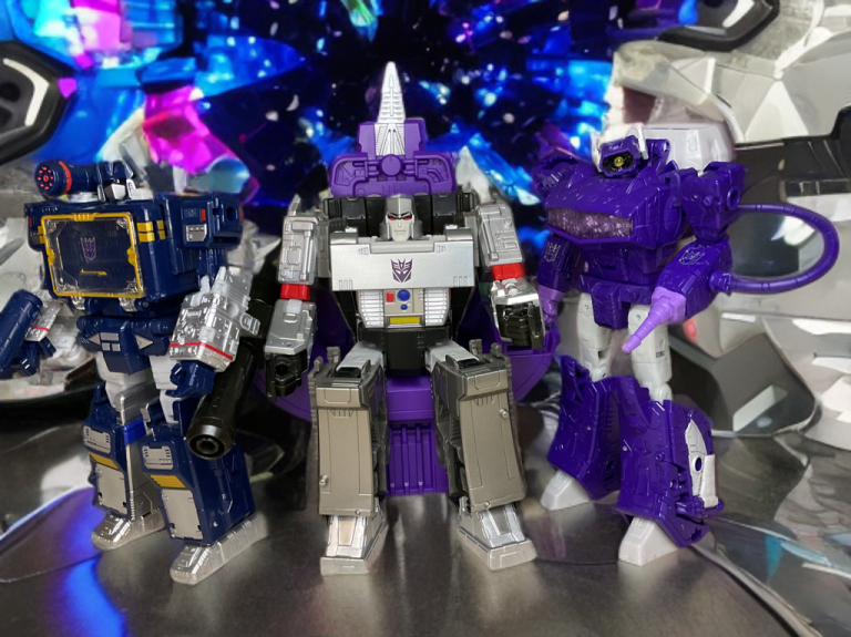 Three Transformers toys: blue Soundwave on left, silver Megatron seated on purple throne in center, purple Shockwave on right, against a starry blue background.